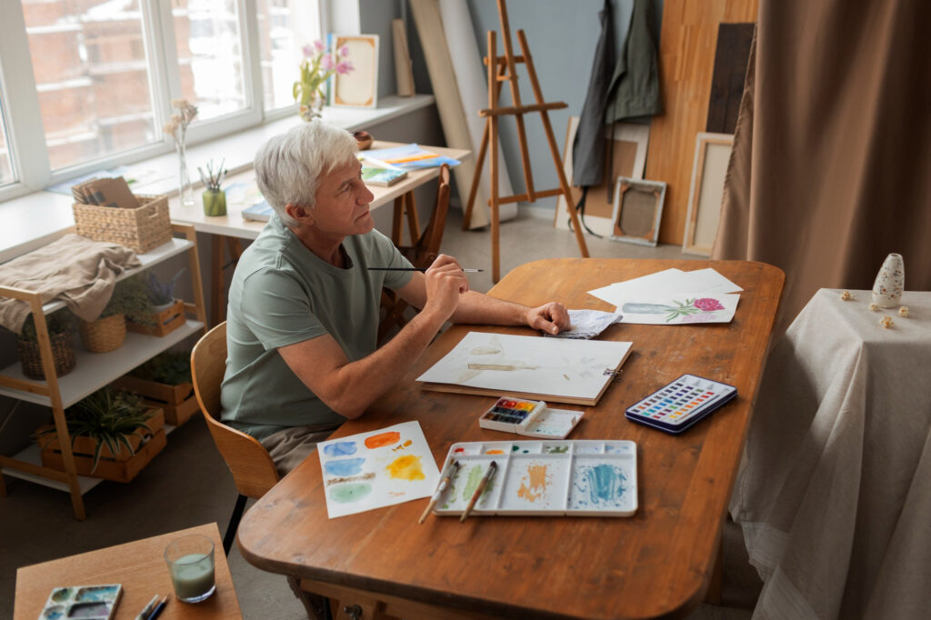 Un homme âgé assis dans un atelier, en train de peindre à l’aquarelle